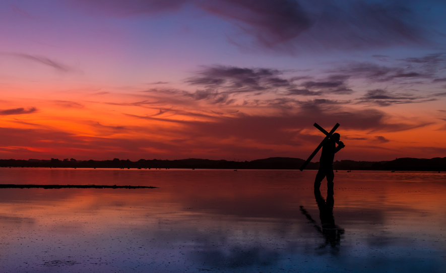 Man carrying a cross on a lake, with a fire like sunset in the sky.